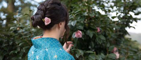 A person in a blue patterned garment standing in front of a bush with pink flowers.