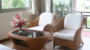 Um conjunto de cadeiras e sofá de vime, uma mesa de vidro, um vaso com flores e vista para palmeiras através de uma janela.