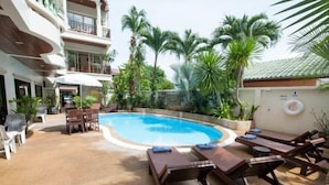 Uma área de piscina com espreguiçadeiras e uma mesa, cercada por palmeiras e um edifício com varandas.