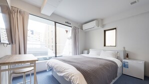 Habitación de hotel con cama, escritorio, silla, ventana con cortinas, aire acondicionado y mesita de noche con una botella.