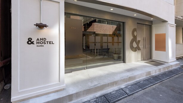 Entrada de hotel con un gran ventanal, un letrero que dice "AND HOSTEL KANDA" y una escalera visible que conduce al interior.