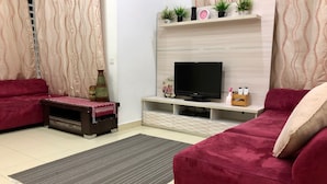 Un soggiorno con televisore su un mobile, due divani rossi, un tavolino con oggetti decorativi e una mensola con piante e un orologio.