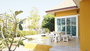 A patio area with a white table and benches, a tiled floor, and a yellow wall. - Baan Tukta (Cha-am)