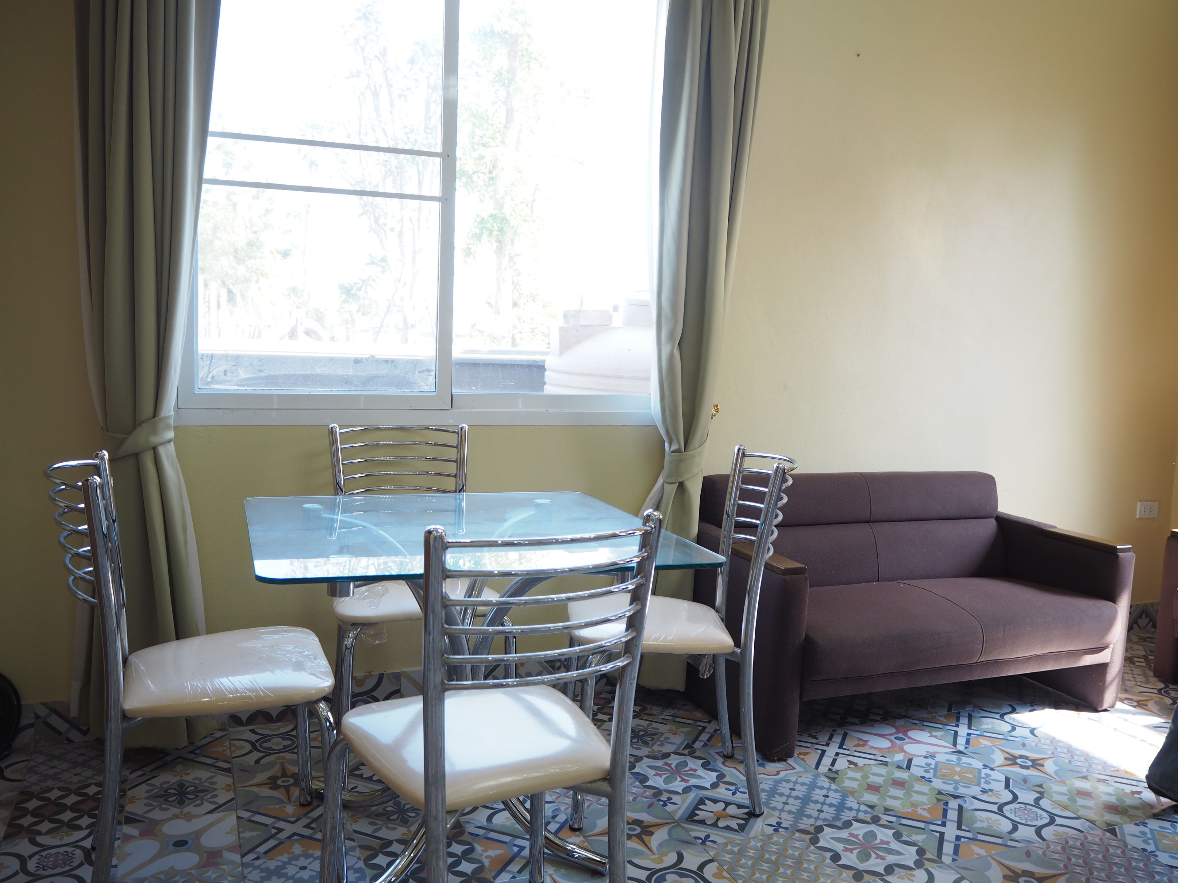 Un comedor con mesa de vidrio y cuatro sillas, un sofá marrón y una ventana con cortinas.
