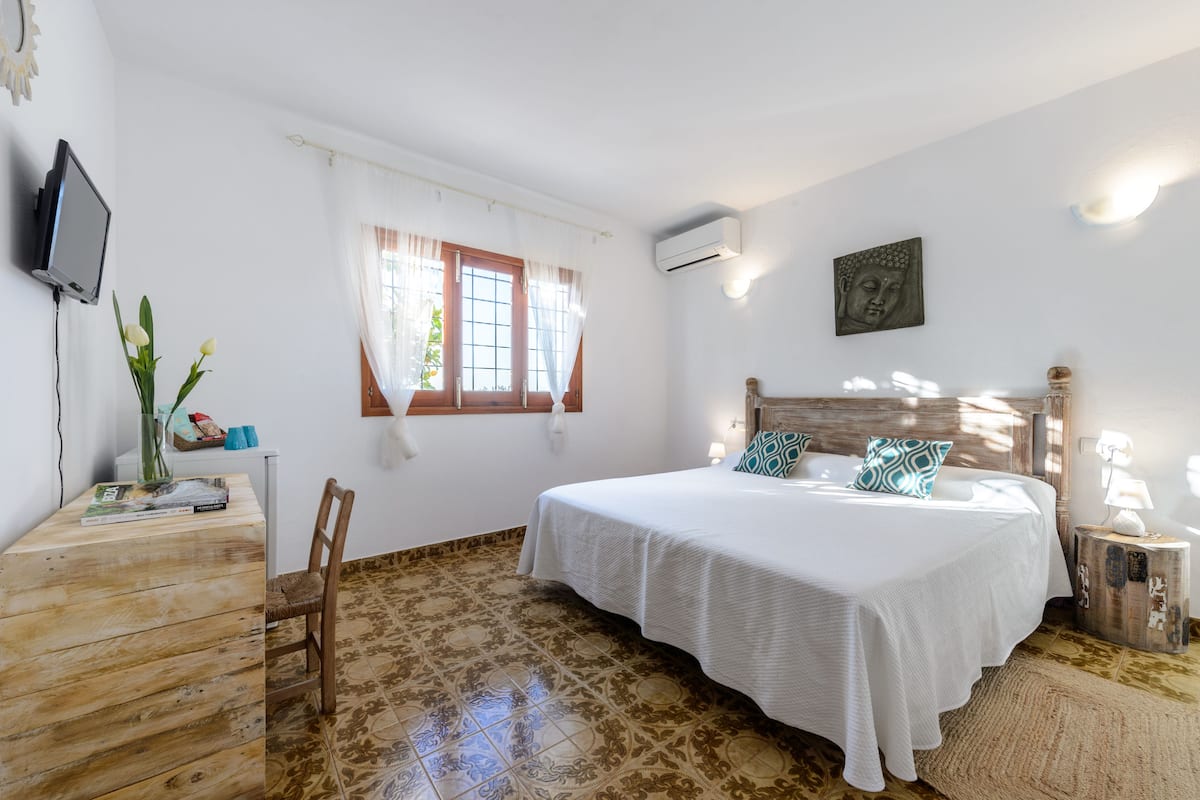 A bedroom with a bed, a desk, a chair, a window, and a Buddha statue.