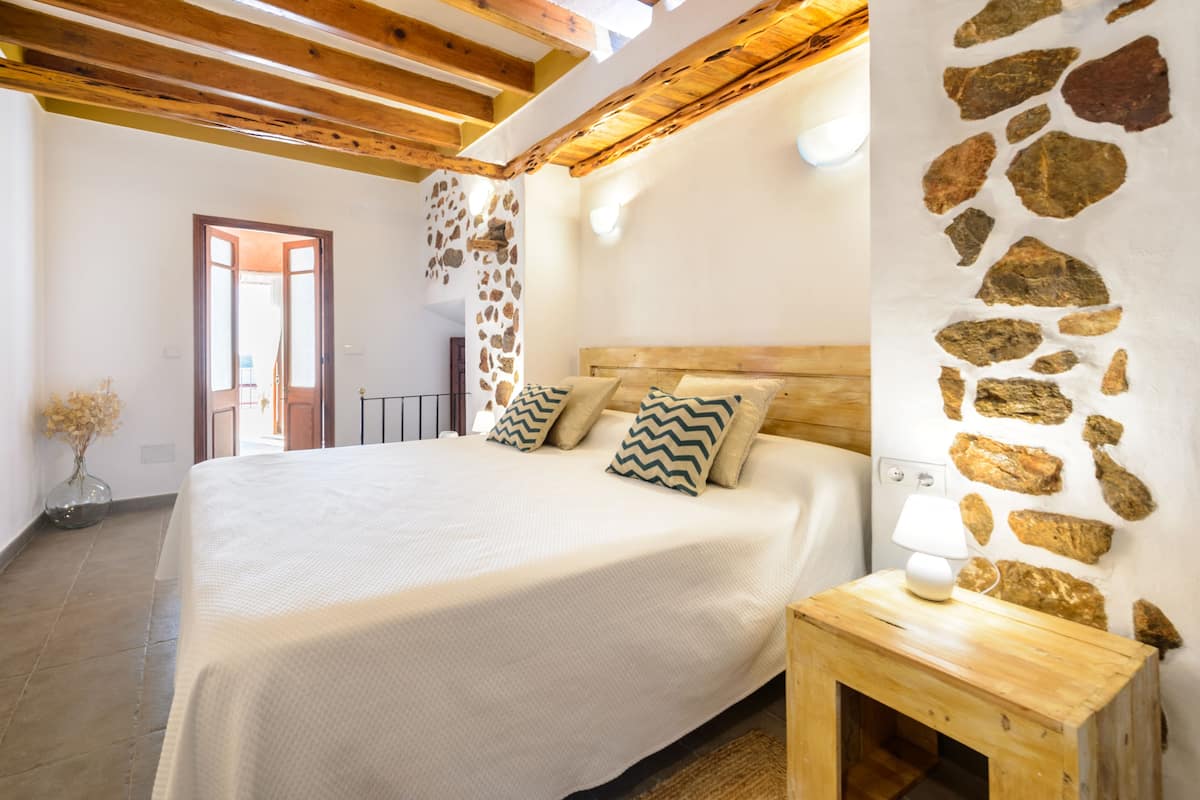 A bedroom with a large bed, wooden beams, stone wall, and a bedside table with a lamp.