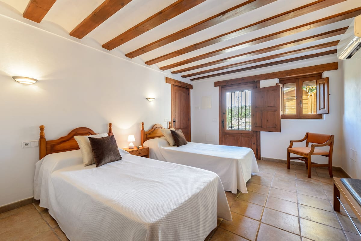 A room with two beds, wooden beams on the ceiling, a wooden chair, and a window with wooden shutters.
