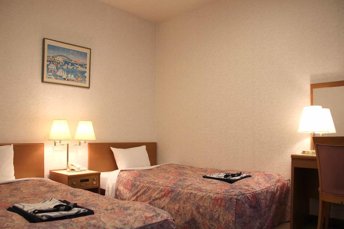 A hotel room with two beds, each with a floral bedspread, a nightstand with a lamp, a chair, and a desk with a lamp.