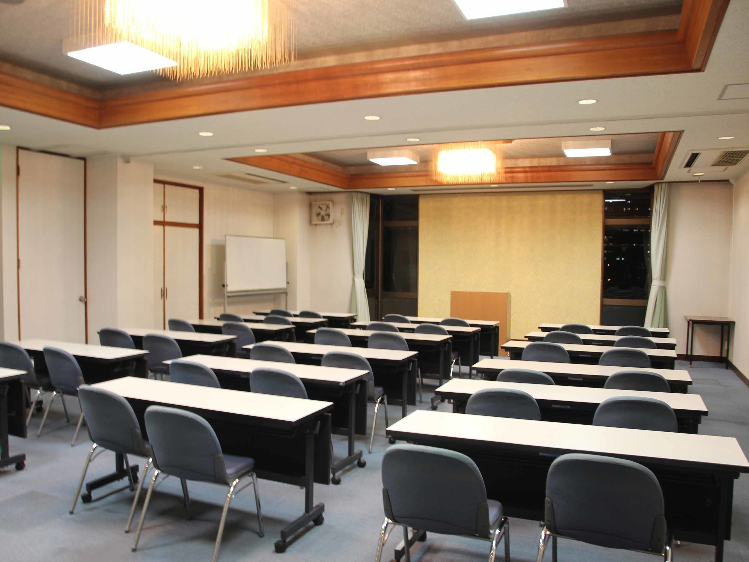 Uma sala de conferências com fileiras de mesas e cadeiras, um quadro branco, um relógio e uma grande janela com cortinas.