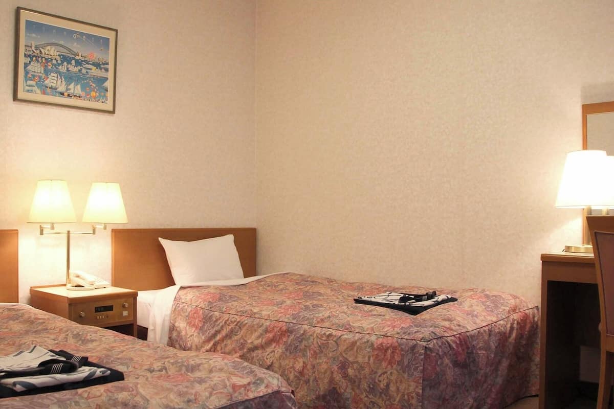 A hotel room with two beds, a nightstand with a lamp, a chair, and a desk with a lamp.