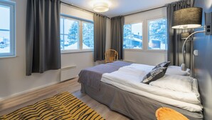 Une chambre moderne avec un grand lit, deux fenêtres avec des rideaux, une chaise et une lampe.