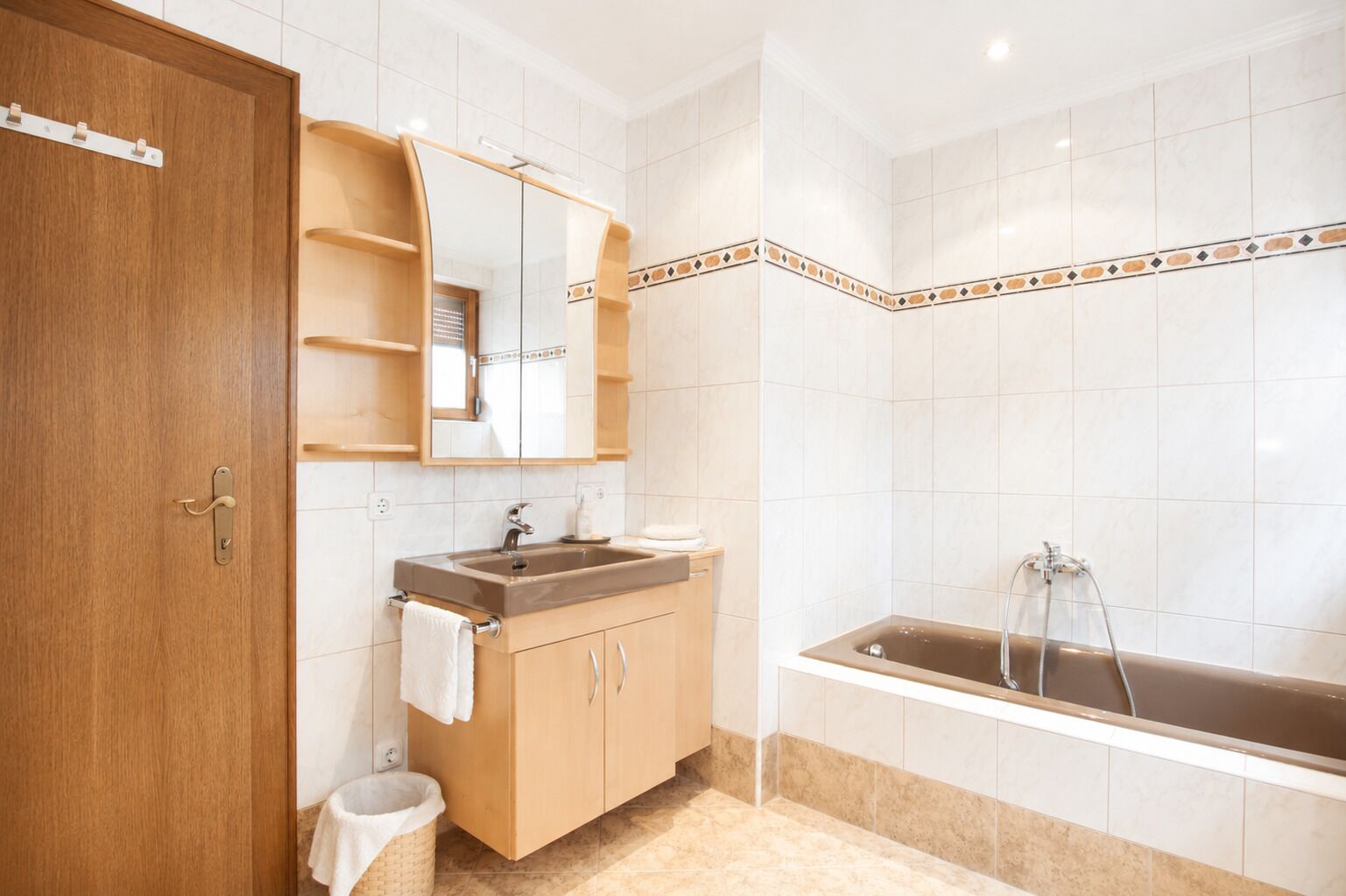 A bathroom with a wooden door, a sink with a mirror, a bathtub, and a tiled wall.