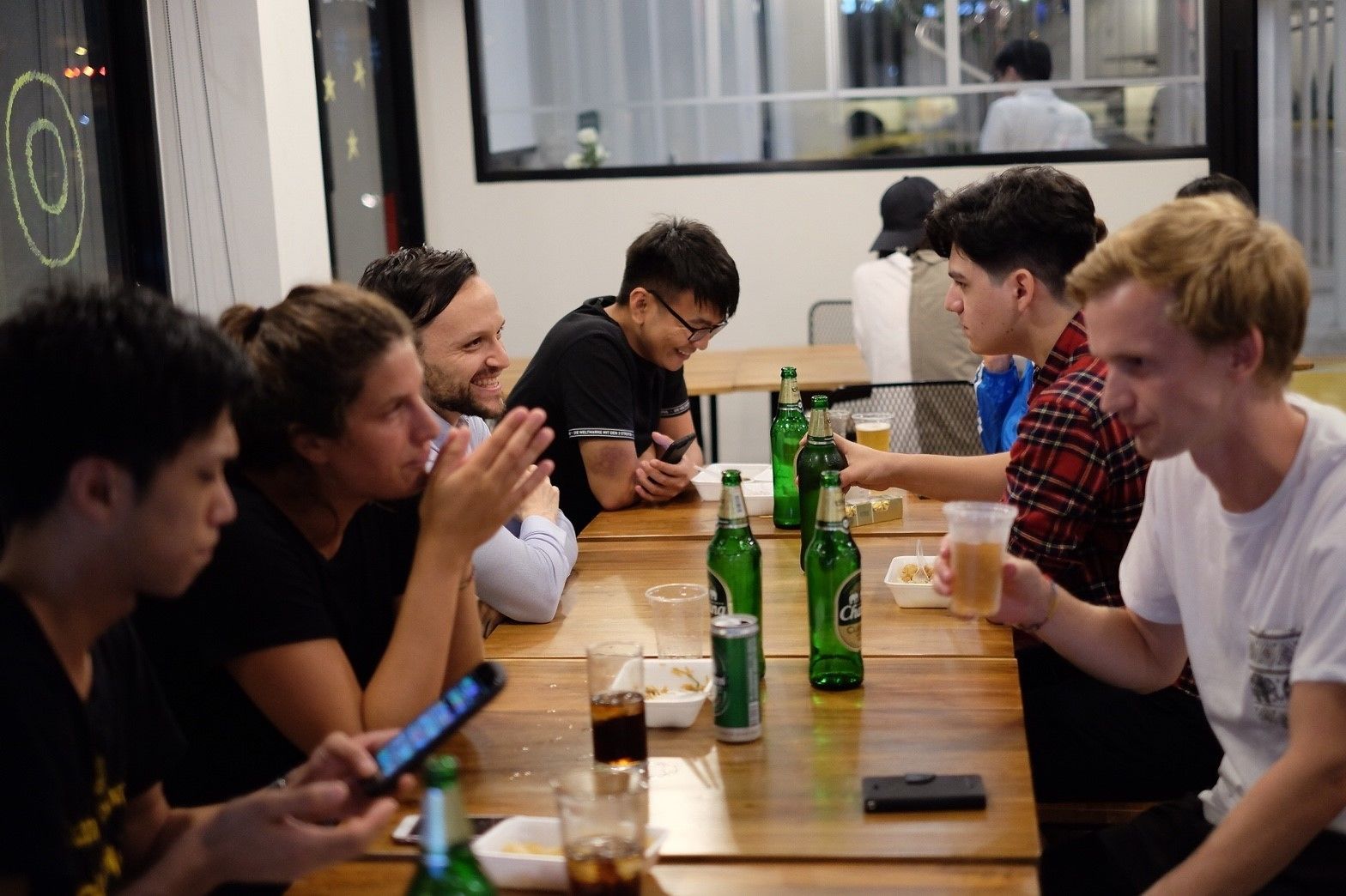 A group of people sitting around a wooden table with green bottles and glasses, in a room with a large window and a view of other people working.