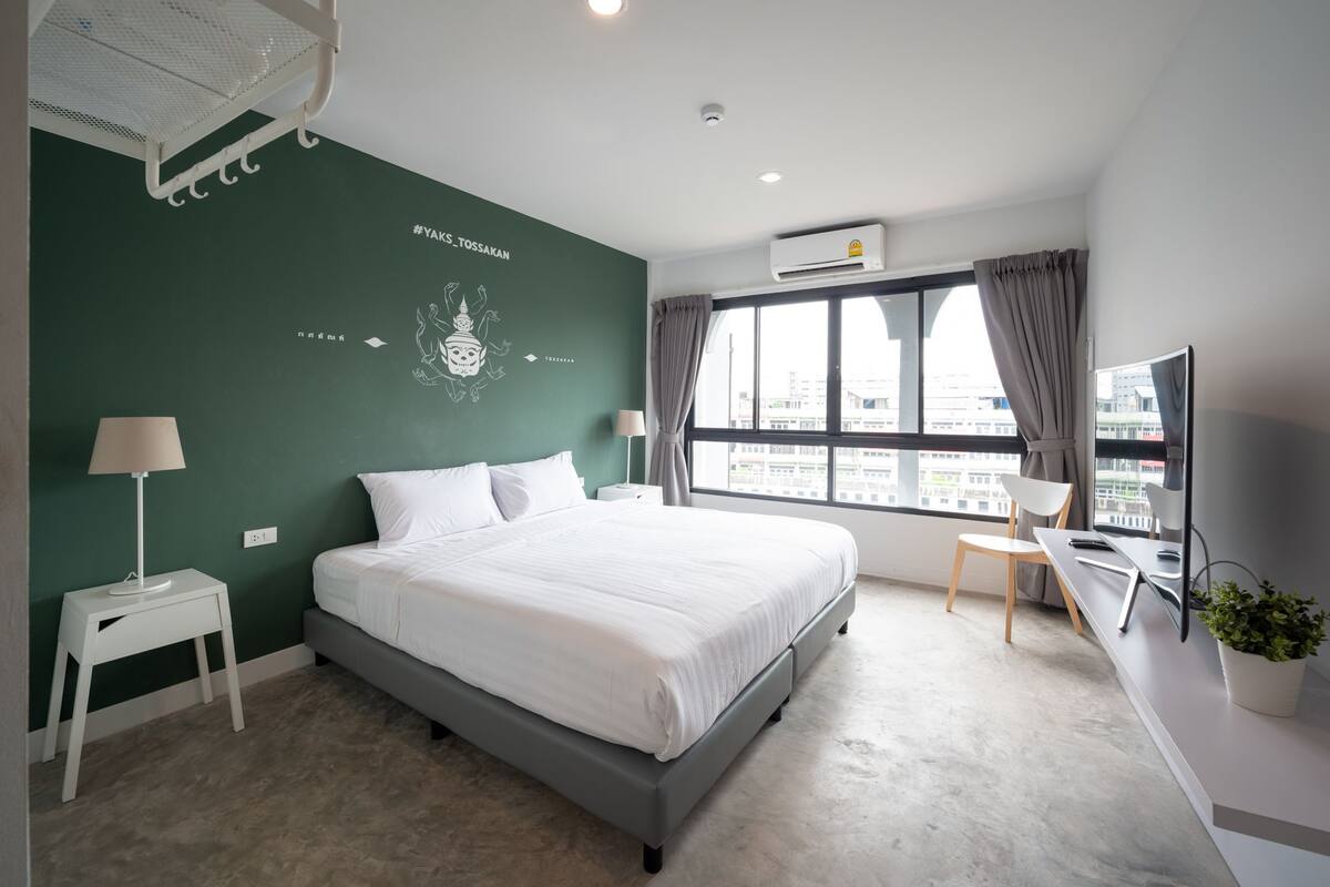 A modern hotel room with a large bed, a desk, a chair, and a window with a city view.