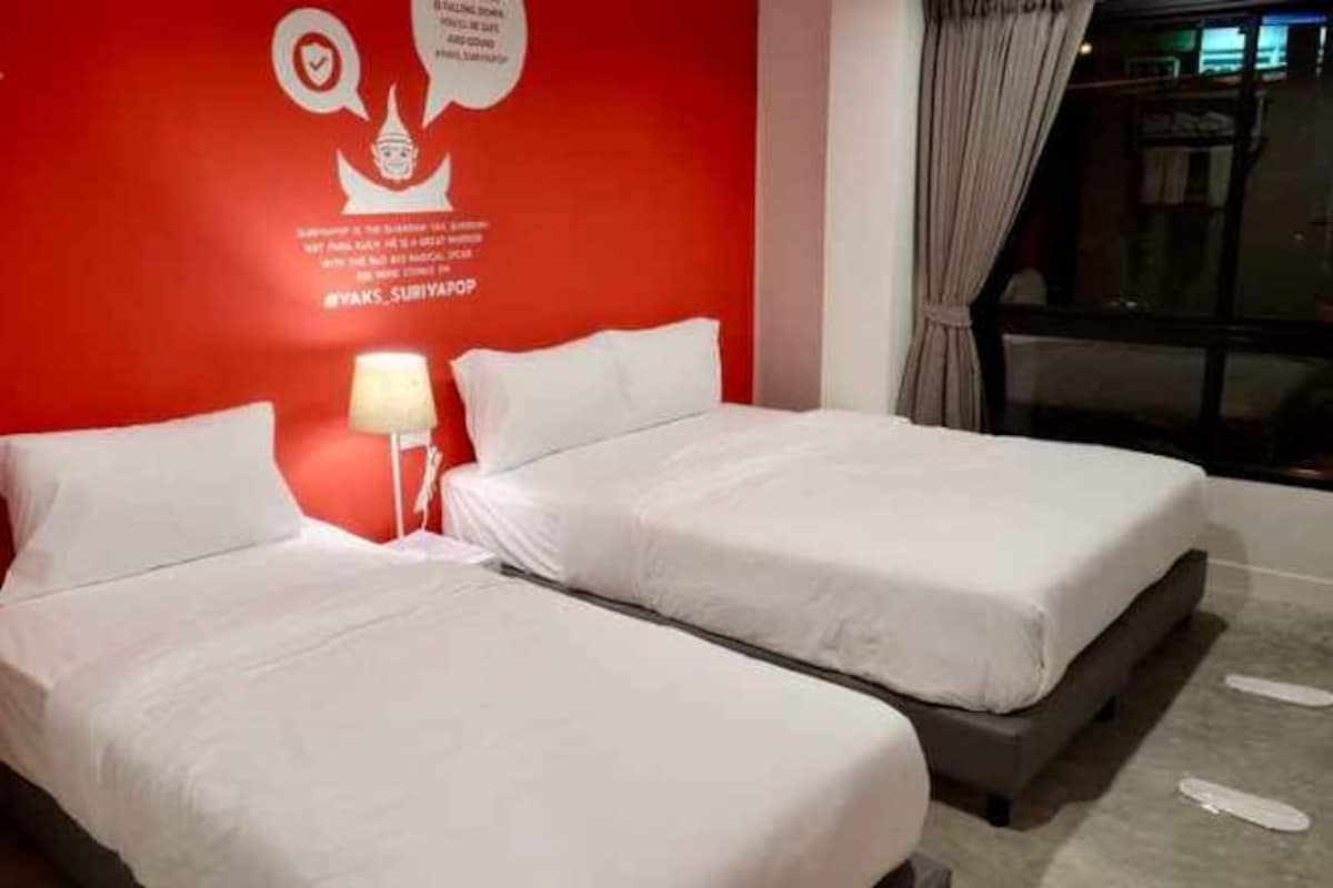 Habitación de hotel con dos camas, una pared de acento roja, una mesita de noche con una lámpara y una ventana con cortinas.