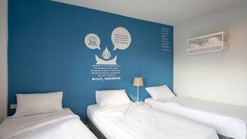 A hotel room with two beds, a blue accent wall, and a shelf with towels.