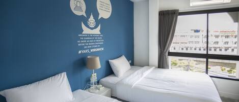 A hotel room with two beds, a blue accent wall, a window with a view of buildings, and an air conditioning unit.