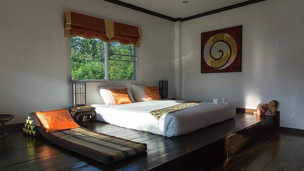 A bedroom with a large bed, a window with a view of greenery, a decorative wall piece, and a wooden platform. - Keawanongburi Resort (Nakhon Ratchasima)