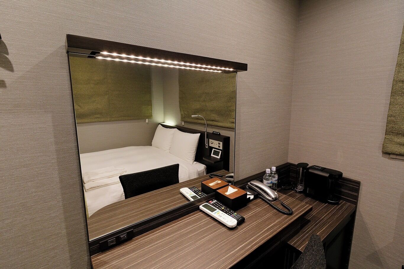 A hotel room with a bed, bedside table with a phone, a mirror, and a desk with a lamp.