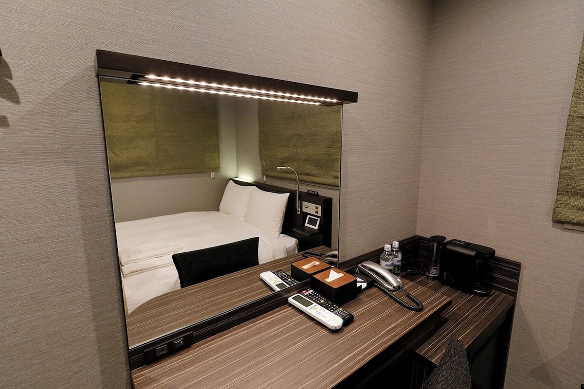 A hotel room with a bed, bedside table with a phone, a mirror, and a desk with a lamp.