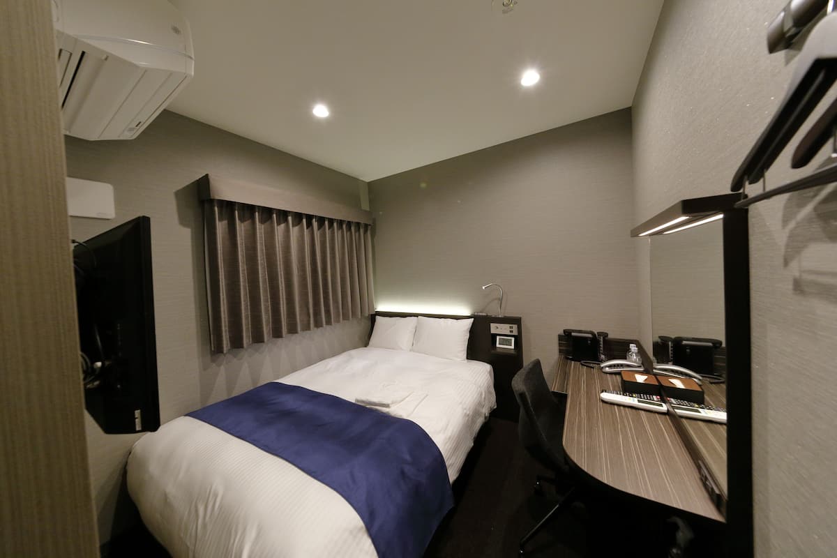 A hotel room with a bed, a desk with a chair, a mirror, a TV, and an air conditioning unit.