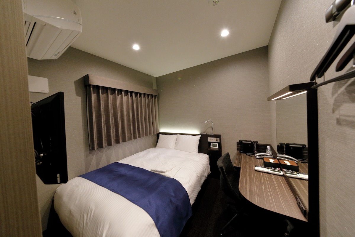 Una camera d'albergo con un letto, una scrivania con sedia, uno specchio, una TV e un condizionatore d'aria.