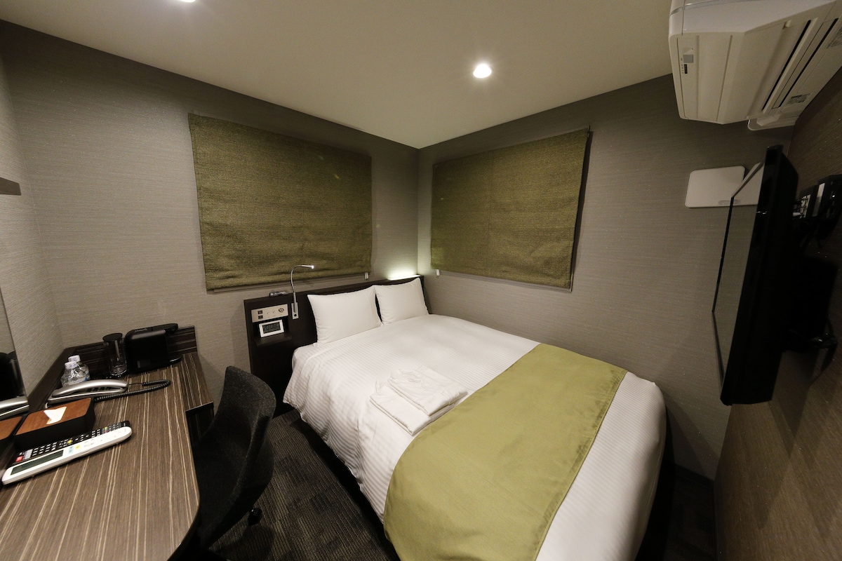 Una habitación de hotel con cama, escritorio con teléfono y control remoto, una silla y aire acondicionado instalado en la pared.