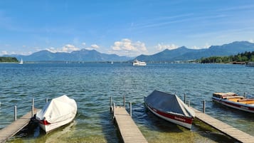 A lake with boats docked at wooden piers, a distant sailboat, and mountains in the background.