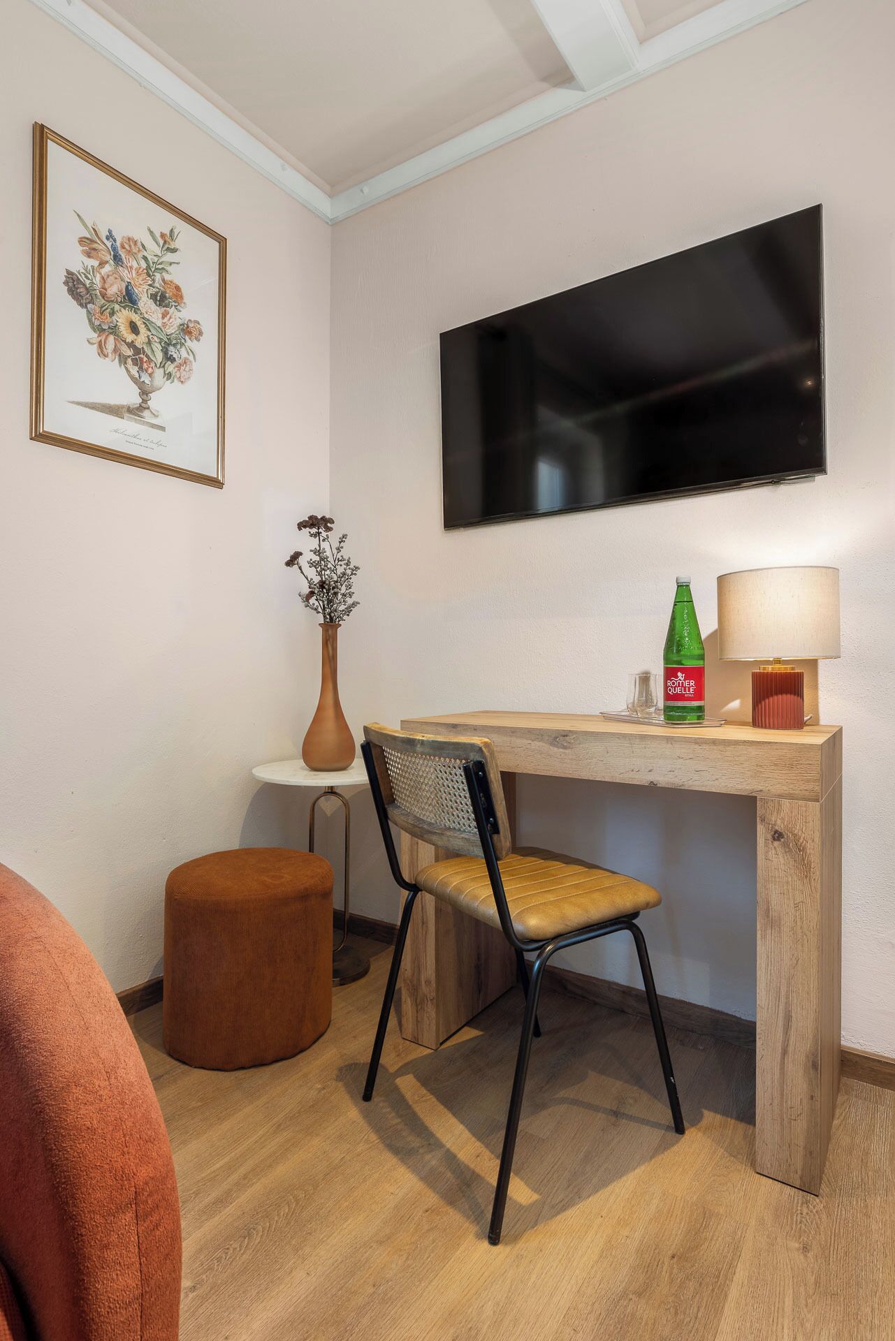 Una habitación pequeña y bien iluminada con un escritorio de madera, una silla, un televisor montado en la pared, una obra de arte floral enmarco, un jarrón con flores secas y una botella de agua.