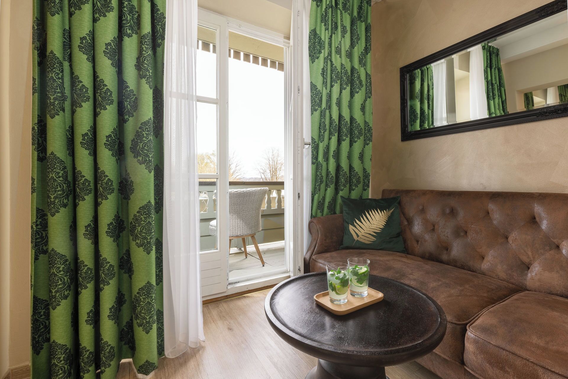 Ein Wohnzimmer mit einem braunen Sofa, einem runden Couchtisch mit zwei Gläsern mit Getränken und grün gemusterten Vorhängen.