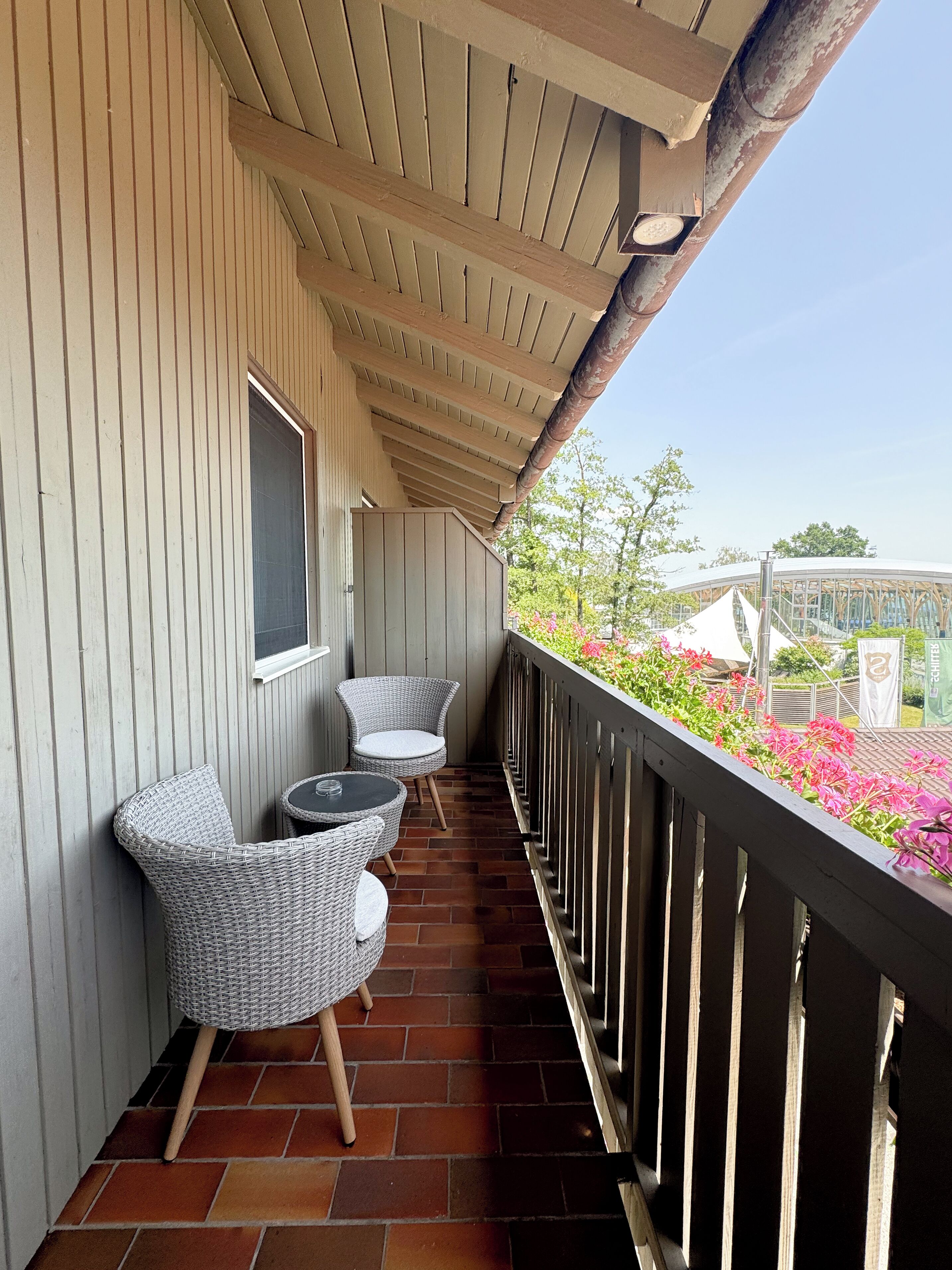 A covered balcony with wicker chairs and a table, a window, and a view of a garden with flowers and a building in the background.
