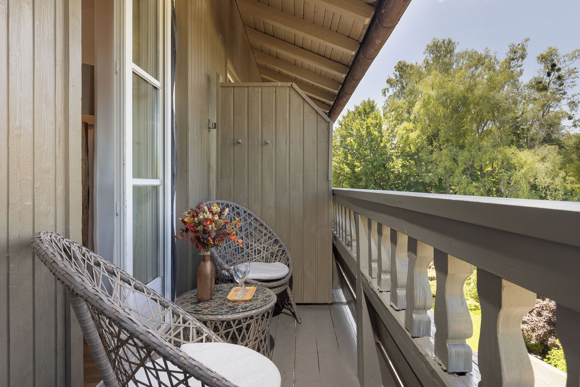 Ein Holzbalkon mit Korbstühlen und einem Tisch, eine Vase mit Blumen und Blick auf Bäume.