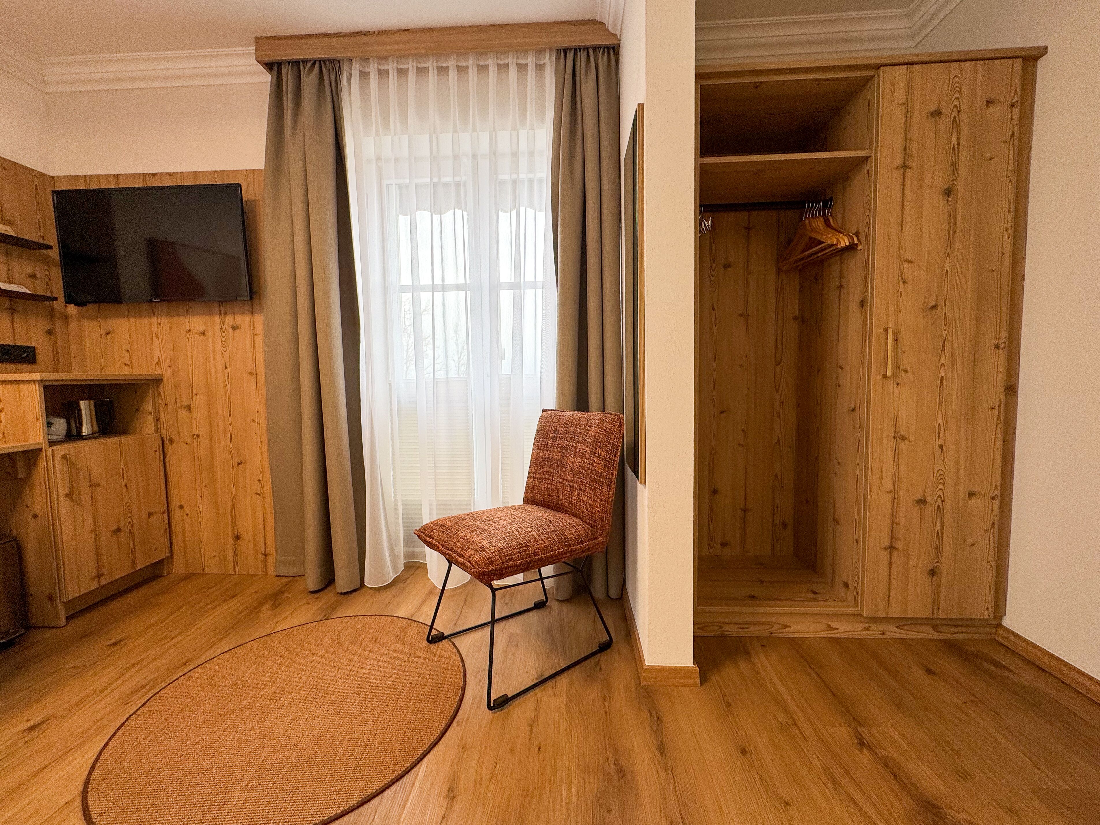 A room with a wooden floor, a chair, a television, a window with curtains, and a wooden wardrobe.