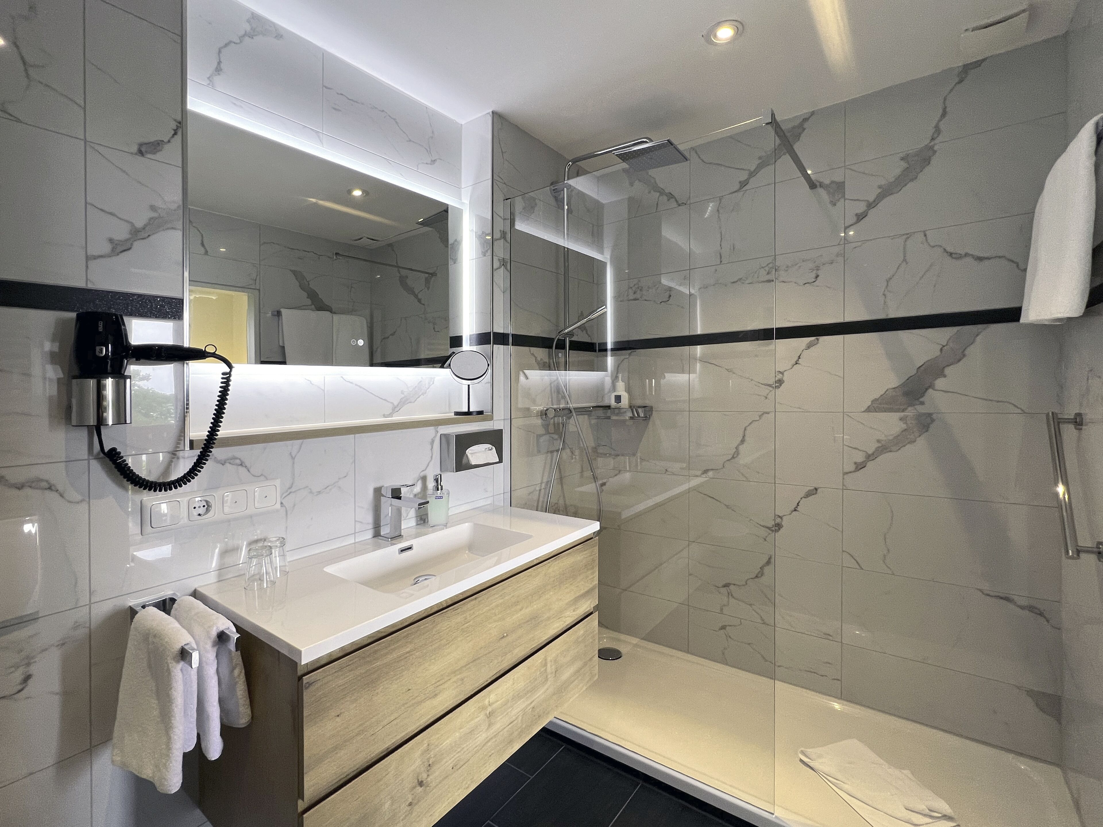 A modern bathroom with a marble backsplash, a glass-enclosed shower, a vanity with a mirror, and a hairdryer mounted on the wall.