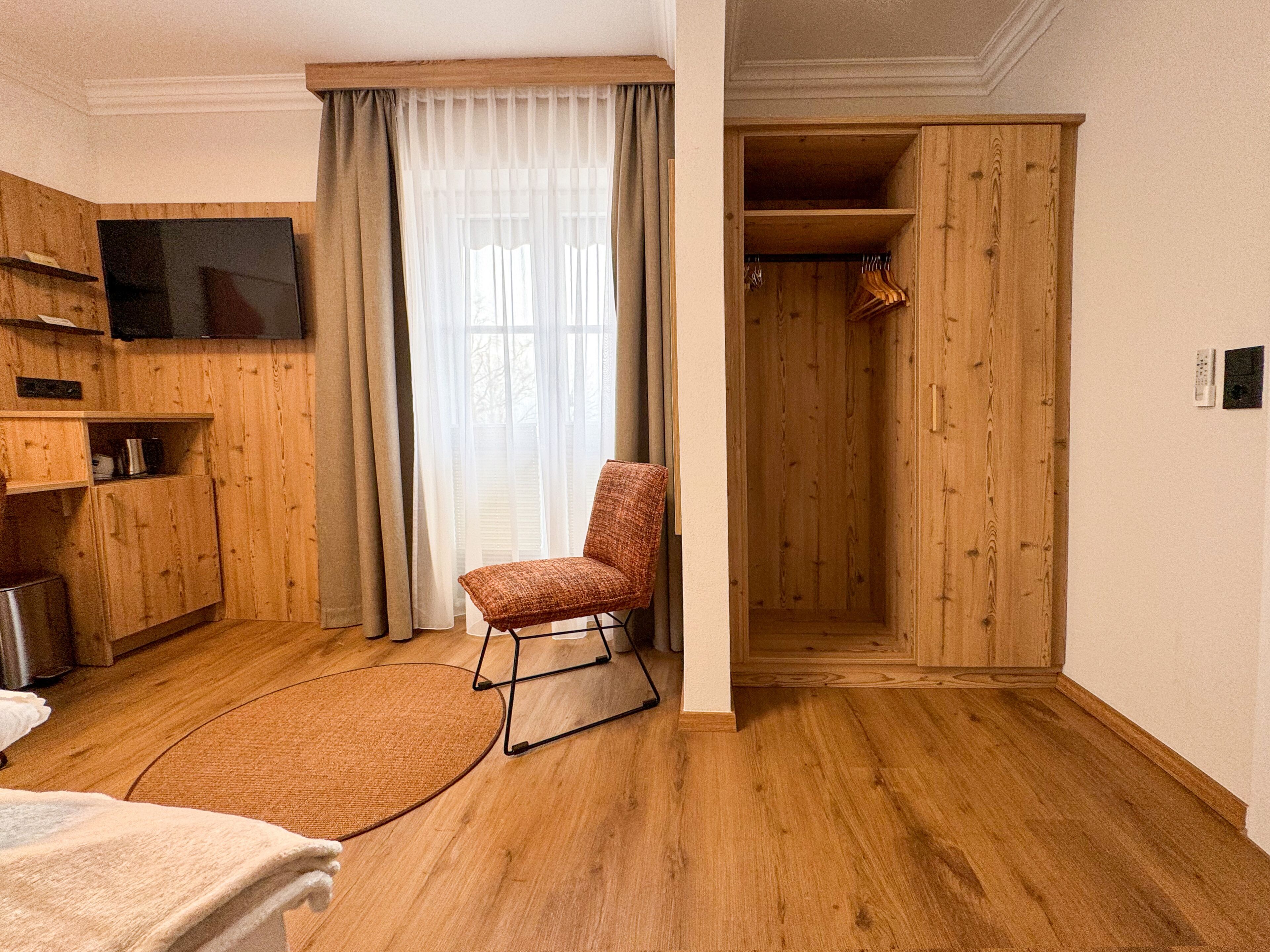 A hotel room with wooden flooring, a television mounted on the wall, a chair, a bed, a wardrobe, and a window with curtains.