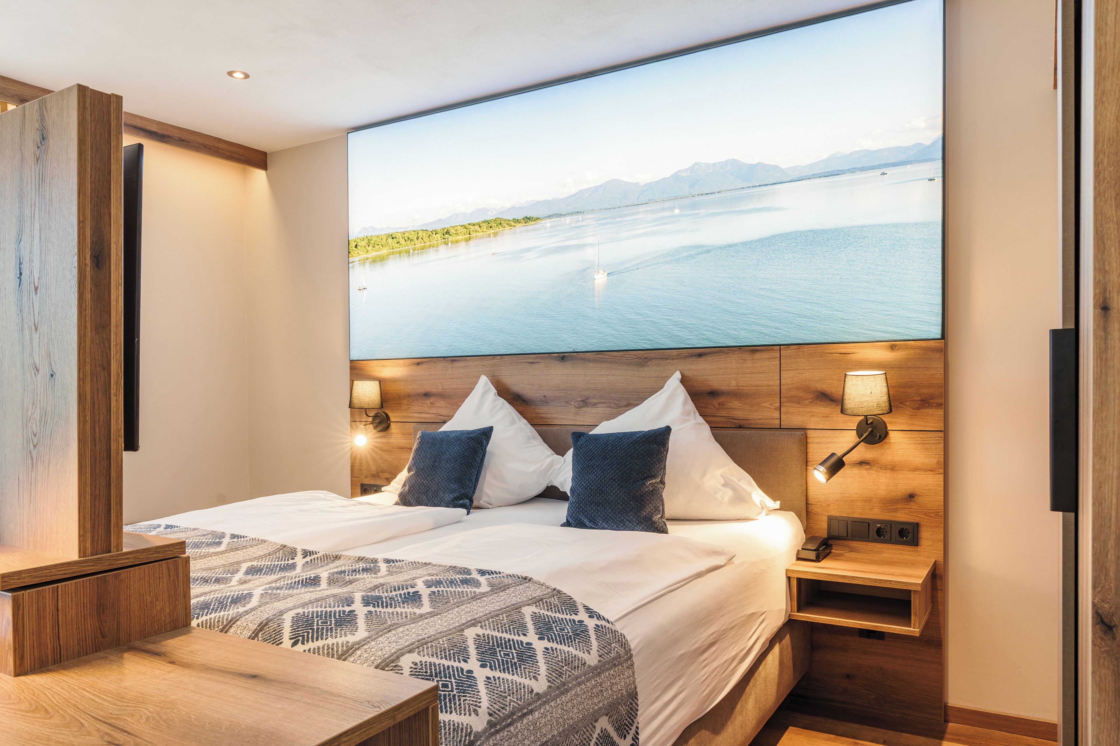 Habitación de hotel moderna con una cama grande, dos mesitas de noche, un velador, un televisor montado en la pared y vistas a un lago y montañas.