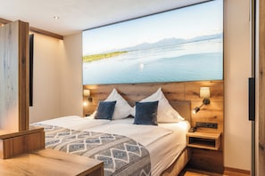 A modern hotel room with a large bed, two bedside tables, a nightstand, a wall-mounted TV, and a view of a lake and mountains. - Hotel Schlossblick Chiemsee (Prien am Chiemsee)