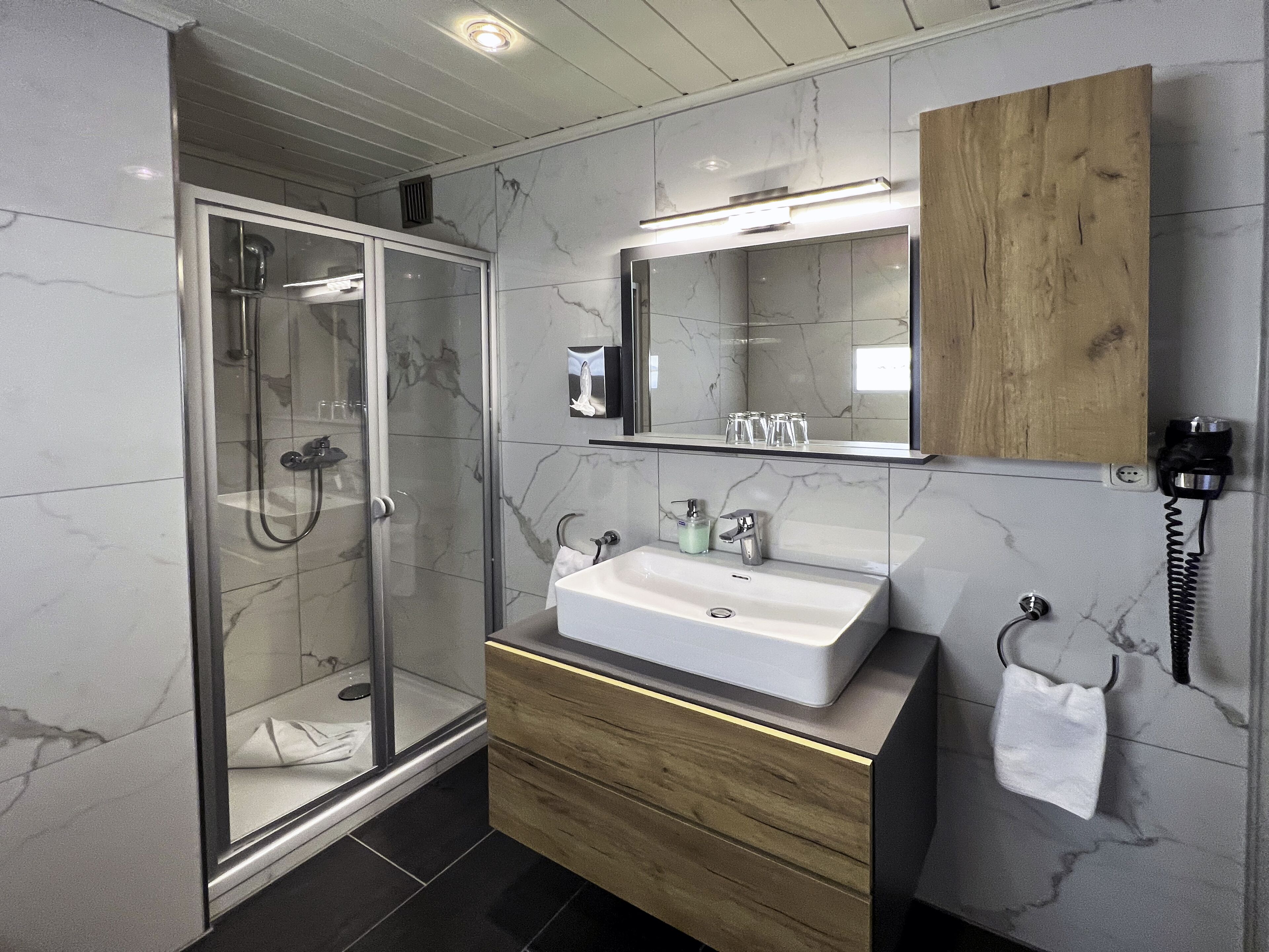 A modern bathroom with a glass-enclosed shower, a wooden vanity, and a marble-tiled wall.