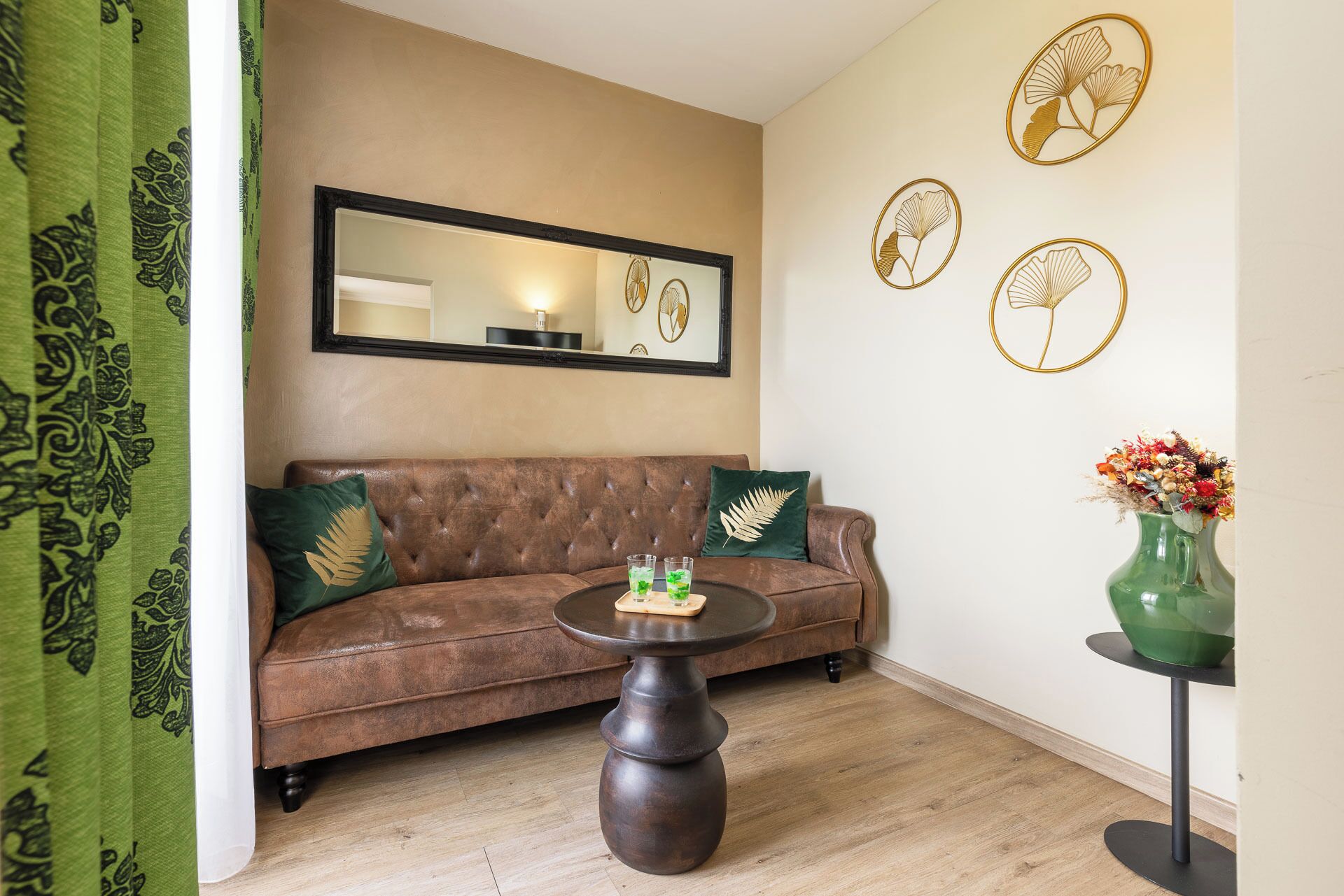 A living room with a brown sofa, a round table with two glasses of water, a green vase with flowers, and decorative wall art.