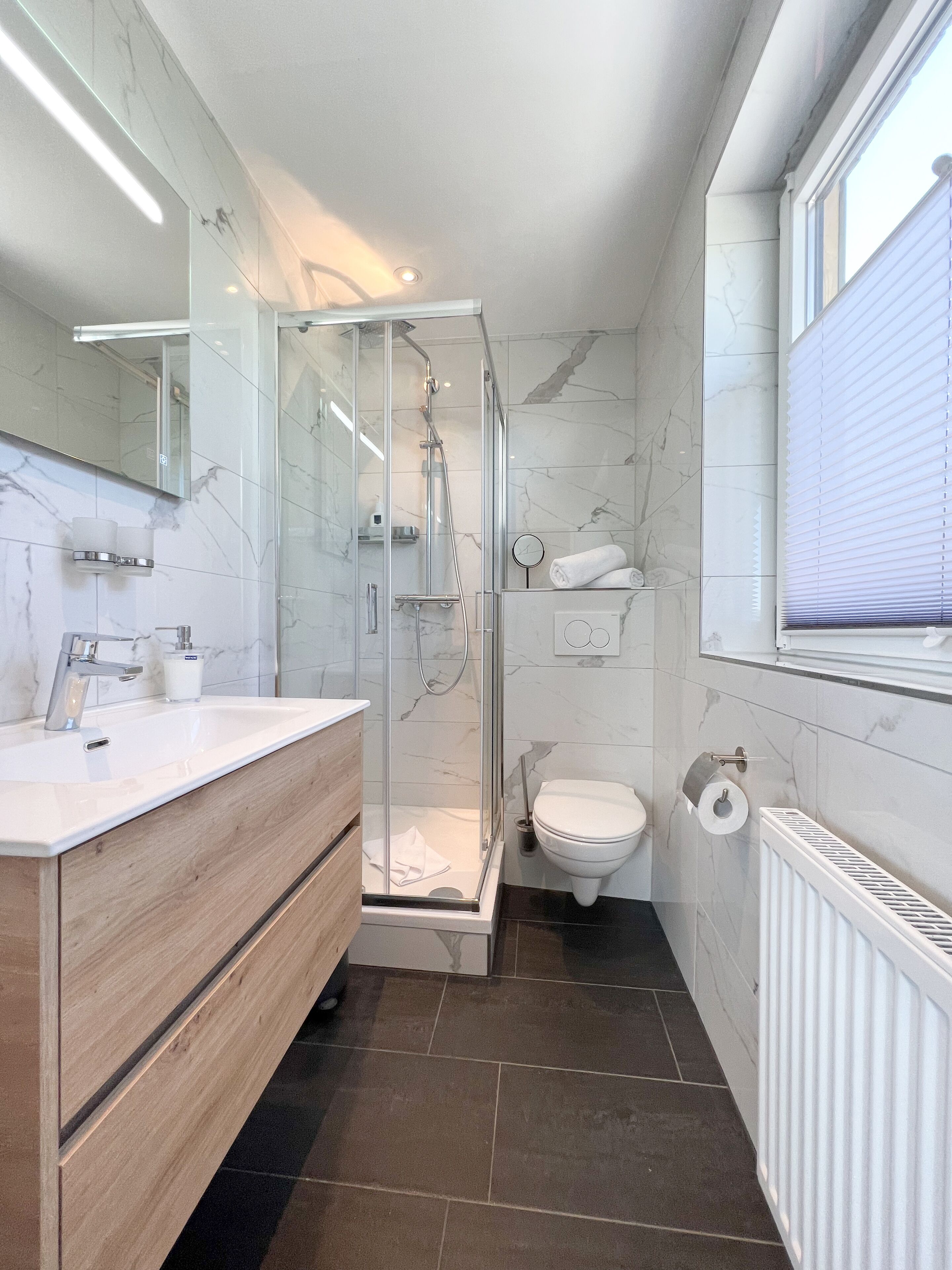 A modern bathroom with a glass-enclosed shower, a wooden vanity, a toilet, and a window with blinds.
