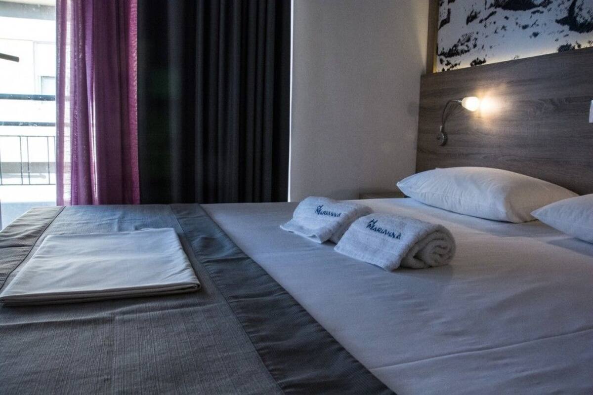 Ein ordentlich bezogenes Bett mit weißen Leinen und zwei Handtüchern mit dem Namen "Mariana", ein Kopfteil mit einer Leuchte sowie ein Fenster mit Vorhängen.