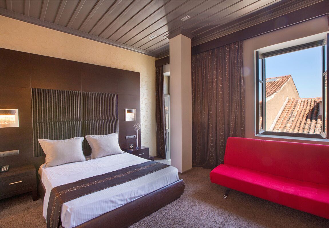 A hotel room with a large bed, a red sofa, a window with a view of buildings, and a wooden ceiling.