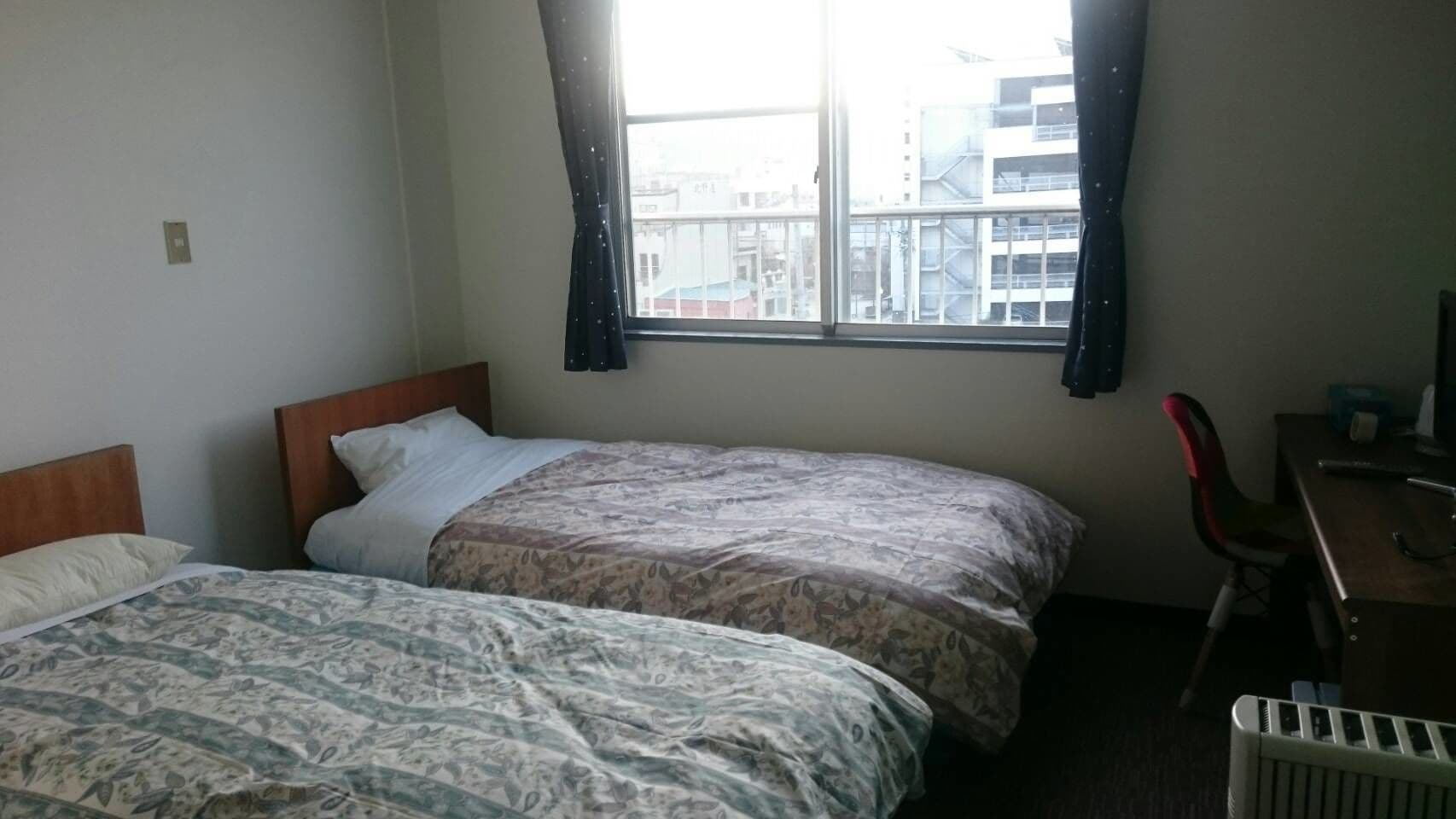 Ein Hotelzimmer mit zwei Betten, einem Schreibtisch, einem Sessel und einem Fenster mit Blick auf die Stadt.