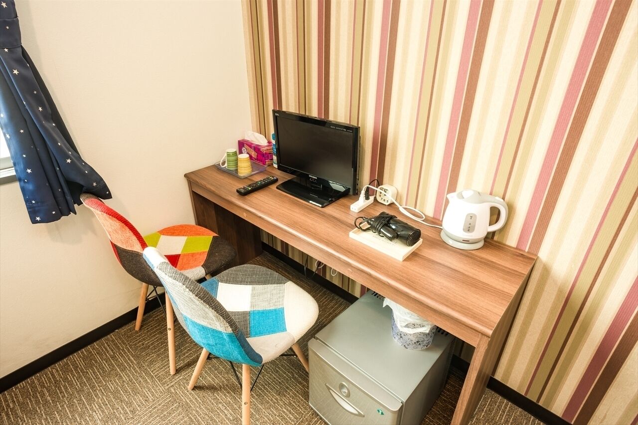 A hotel room with a wooden desk, a black monitor, a remote control, a camera, and a kettle. There is a colorful chair and a small filing cabinet.