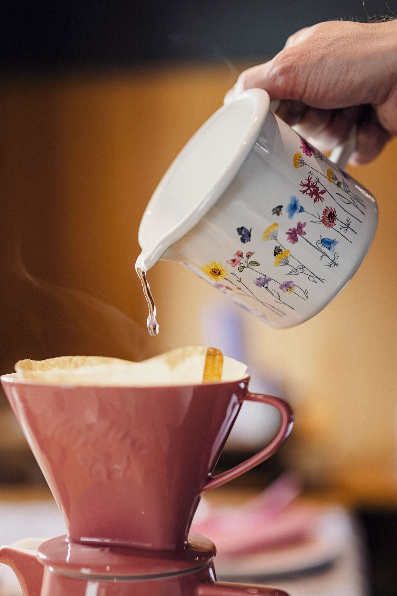 A hand pouring liquid from a floral-patterned mug into a red cup.