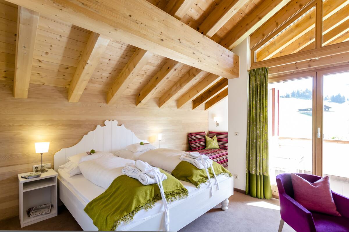 Un dormitorio con techo de madera, una cama con ropa de cama blanca, un plaid verde, una mesita de noche y una silla.