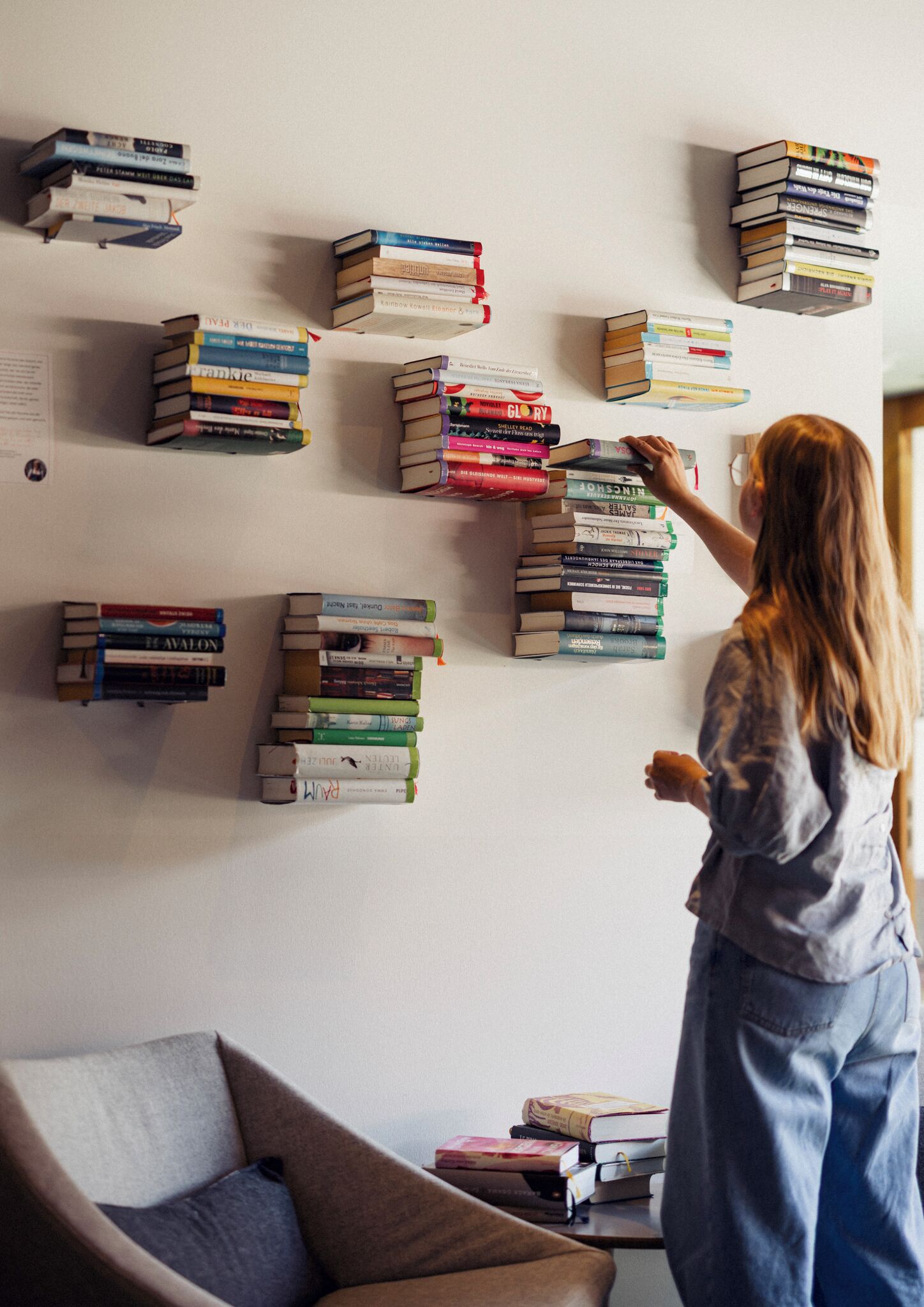 A person arranging books on a wall-mounted shelf in a room with a couch and a small table.