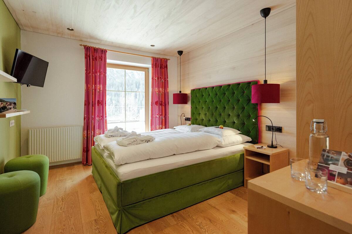 Un dormitorio con una cama verde, una mesita de noche de madera, un televisor montado en la pared y una ventana con cortinas rosas.