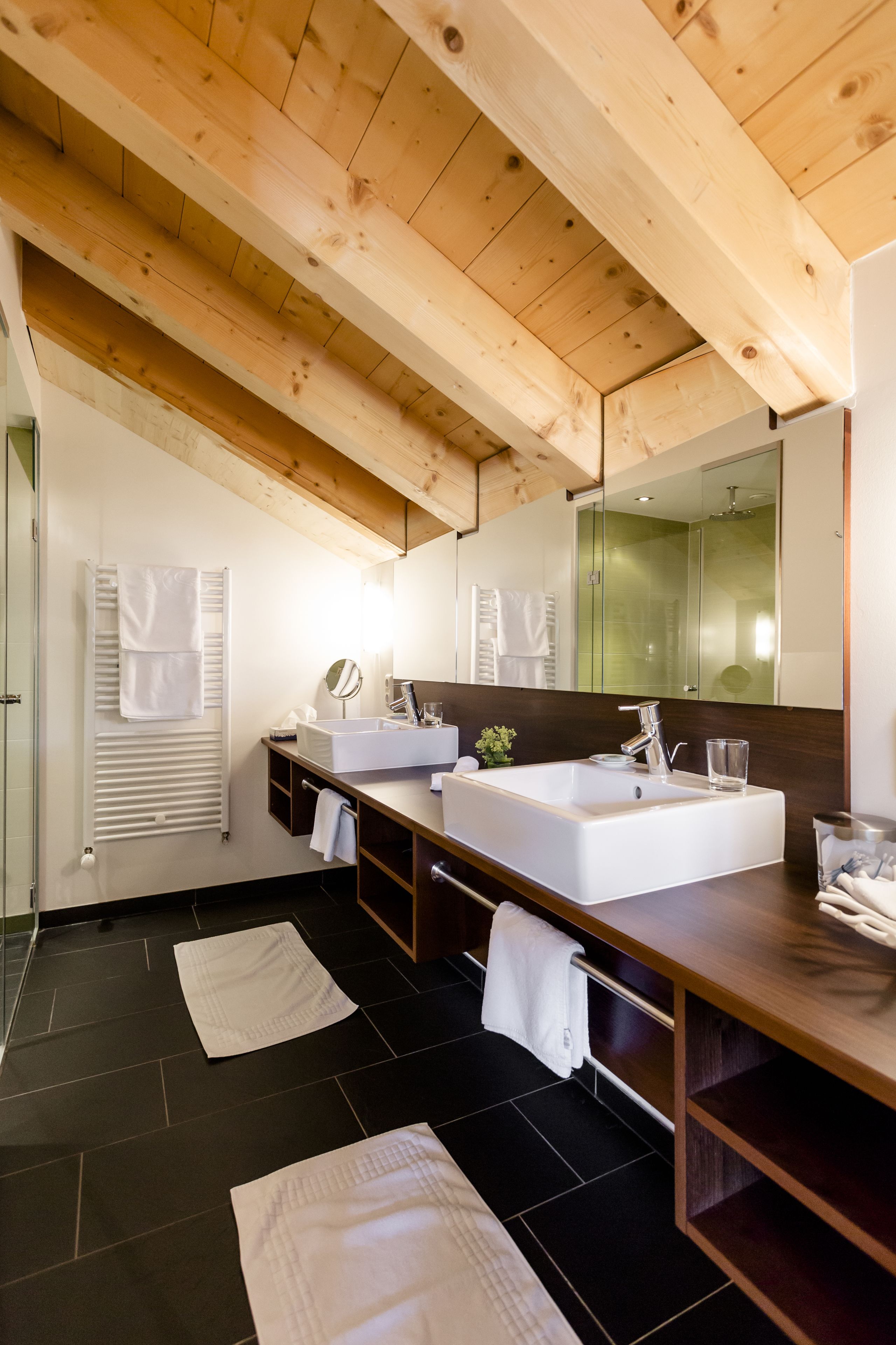 A modern bathroom with a wooden ceiling, a large mirror, two sinks, and black tiled flooring.
