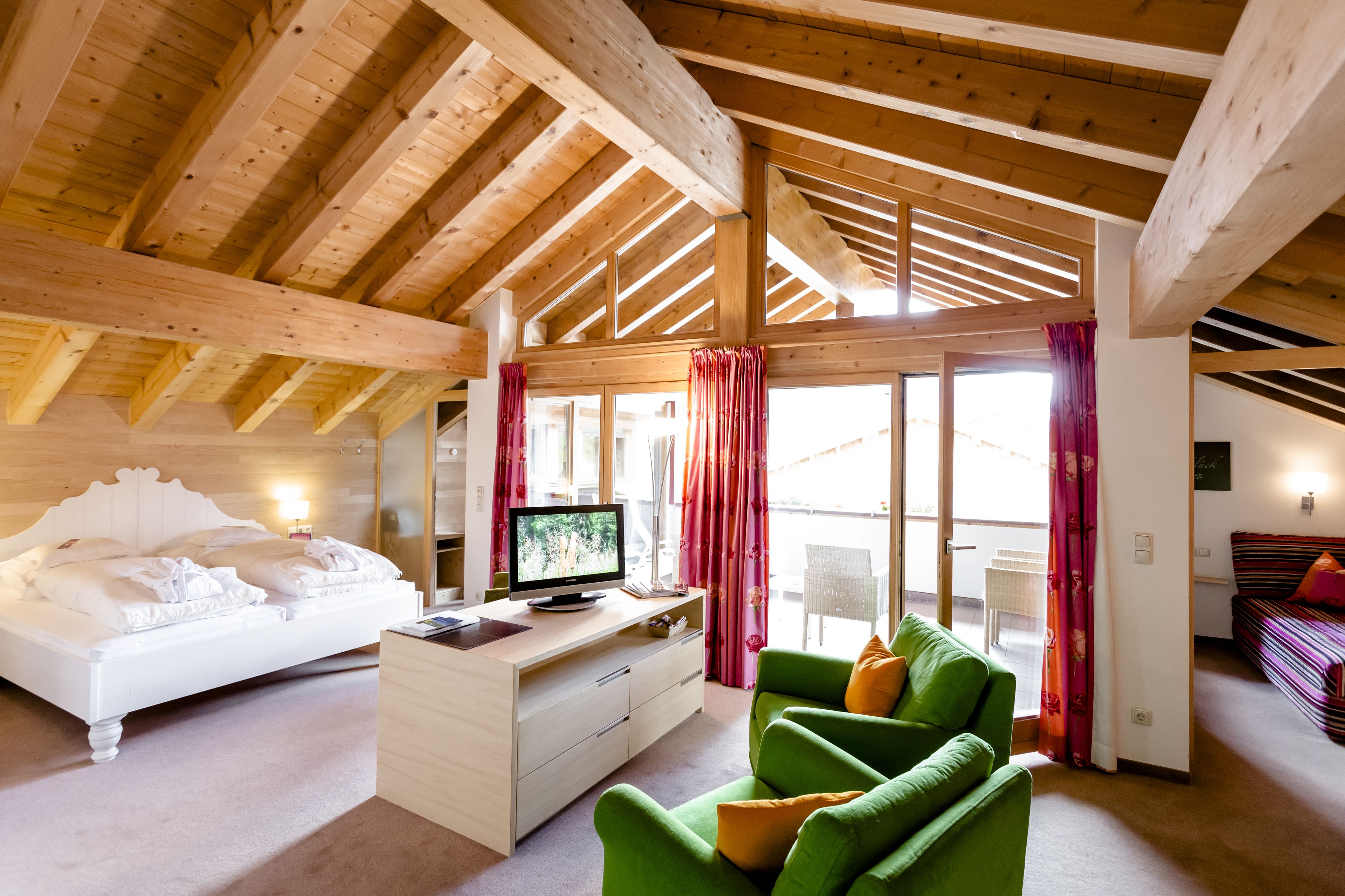 Une pièce spacieuse avec un plafond en bois, un lit recouvert de linge de lit blanc, un canapé vert et un téléviseur posé sur une commode en bois.